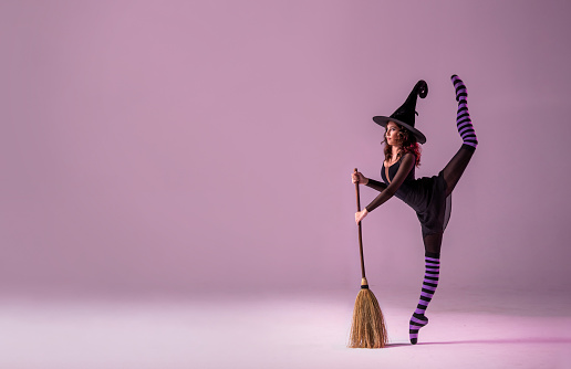 ballerina on pointe shoes in a black witch costume in hat and with broom is dancing on roof on lilac background.