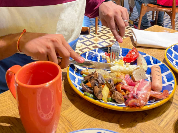 istock Image of unrecognisable, Indian Hindu man cutting into fried egg with knife and fork, eating breakfast in hotel restaurant, fry up breakfast with sausages, bacon, fried mushrooms and tomato, blurred diners background, focus on foreground 1427004424