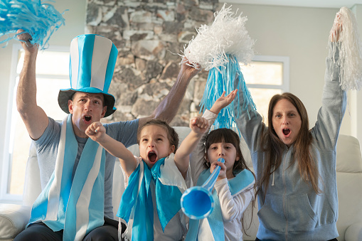 Portrait of a soccer fan argentine family (father, mother and two daughters) watching a soccer match on tv at their home and uproariously celebrating a goal. They are wearing and using merchandising (pom poms, vuvuzela, flag and hat).