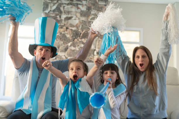 Portrait of soccer fan argentine family happily  watching soccer match on tv at home and celebrating a goal Portrait of a soccer fan argentine family (father, mother and two daughters) watching a soccer match on tv at their home and uproariously celebrating a goal. They are wearing and using merchandising (pom poms, vuvuzela, flag and hat). argentina stock pictures, royalty-free photos & images