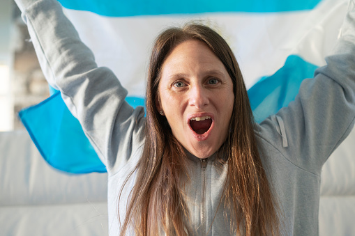 Close-up  of  argentine soccer female fan with a flag watching a soccer match on tv and celebrating a goal. She is sitting on a couch.