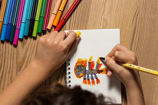top view of a child's hands painting a colorful dragon on a white sheet. The table has colored markers and drawing material