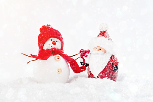 Decorative Santa with a gift stands in the snow with a snowman in a red hat and a red scarf on a light background. Glitter background.New Year's card background.
