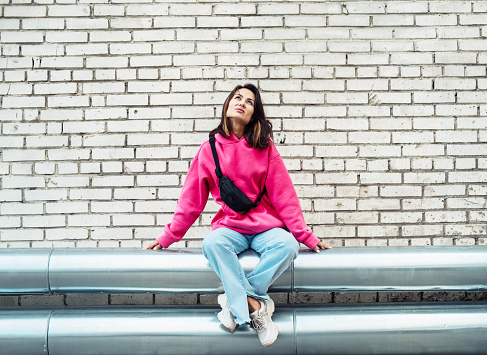 Fashion Asian young woman in a pink sweatshirt and jeans sits against a brick wall background, has fun. The concept of youth and fashion.