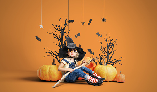 A child girl in a large witch's hat and with a broom in her hands against the backdrop of the scenery for the Halloween holiday. Studio portrait.