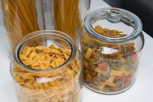 Variety of types and shapes of Italian pasta in glass jars on marble background.
