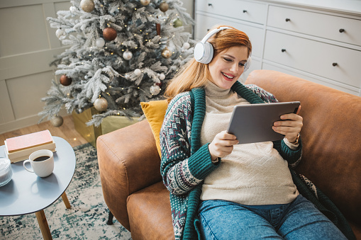 Young pregnant woman at home for Christmas. She is sitting on sofa and using digital tablet for business or watching movies.