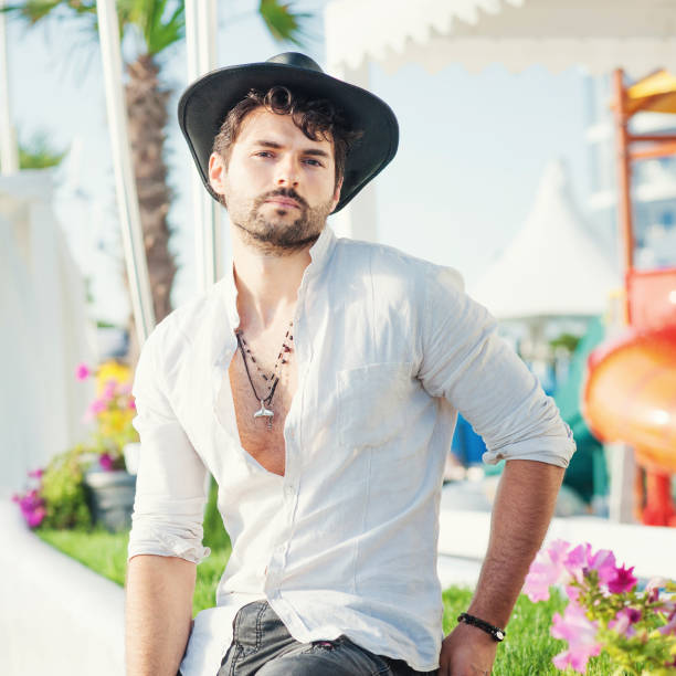Sexy man model dressed in black hat and white shirt, outdoor photo against touristy tropical city landscape