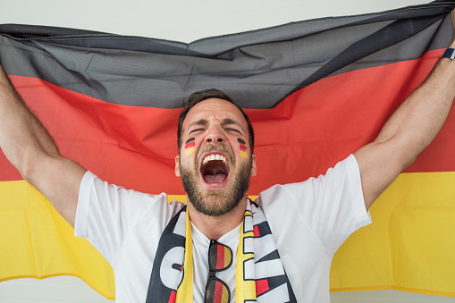 Young man soccer fan cheering at the game. Holding national team flag, with color paint on face. Studio shot