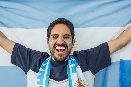 Young man soccer fan cheering at the game. Holding national team flag, with color paint on face. Studio shot