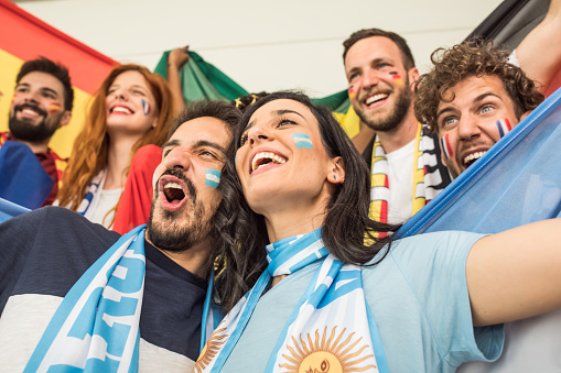 Group of young multi-ethnic soccer fans cheering at the game. Holding national team flags, with color paint on faces. Studio shot