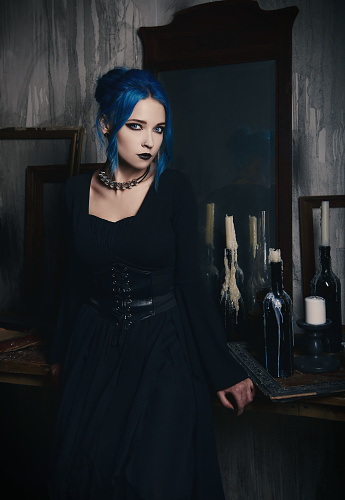 Indoors portrait of gorgeous goth girl in black dress. Blue-haired gothic lady stands at the old mirror. Vintage look