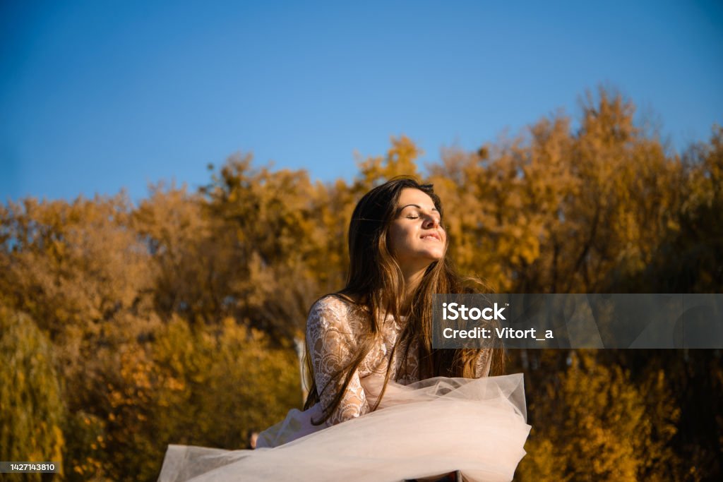 She exposes her face to the autumn sun a girl with long dark hair in a puffy pale pink ball gown stands on a bridge on a clear cloudless day, chiffon fabric flies in a gust of wind. She exposes her face to the autumn sun 25-29 Years Stock Photo
