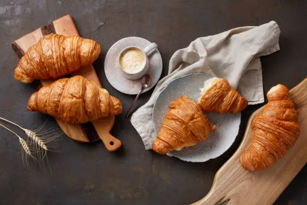 Kitchen boards, gray napkin, plate and cup of coffee with tasty croissants on rusty table. Fresh croissant on brown background with spikelets of wheat. Flat lay. French pastry.