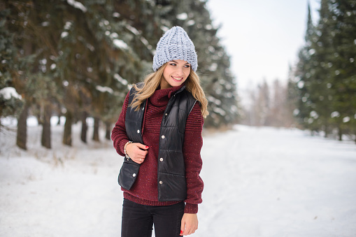 portrait of a happy girl in a blue hat, sweater and vest, stands on a snowy winter alley among the Christmas trees