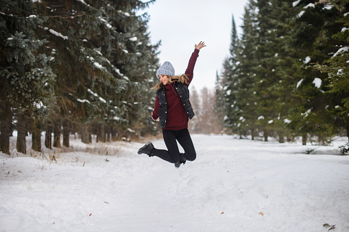 portrait of a happy jumping girl in a blue hat, sweater and vest, on a snowy winter alley among the fir trees
