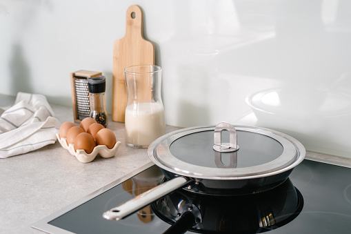stainless steel pan with lid on glass ceramic cooker hob, eggs, milk, pepper and towel on countertop at kitchen with light interior, preparing breakfast at home
