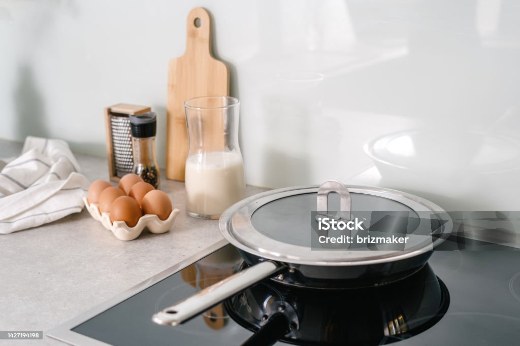 frying pan on electric stove at modern kitchen stainless steel pan with lid on glass ceramic cooker hob, eggs, milk, pepper and towel on countertop at kitchen with light interior, preparing breakfast at home Cooking Pan Stock Photo