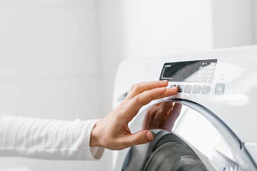 cropped shot of woman hand turn on automatic washing machine or select program with button on control panel in white bathroom in modern house