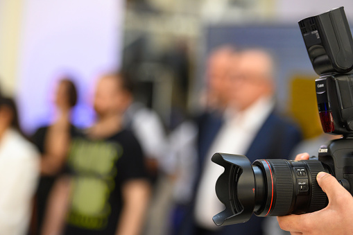 A camera man with two cameras frames his shot for the presentation about to take place on the stage. He is somewhat silhouetted and is wearing headphones.