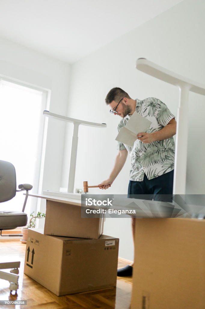A young man using hammer, following instructions and assembling an office desk in his new home Adult Stock Photo