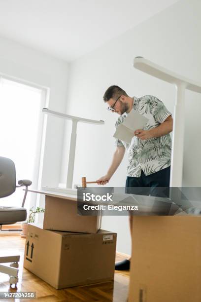 A Young Man Using Hammer Following Instructions And Assembling An Office Desk In His New Home Stock Photo - Download Image Now