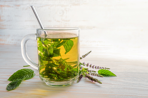 fresh peppermint leaves and flowers brewed as herb tea in a glass cup on a light wooden table, copy space, selected focus, narrow depth of field