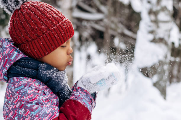 Happy little African-American girl in a red hat and overalls blowing snow off the hand.Winter, Christmas and Happy New Year concept. stock photo