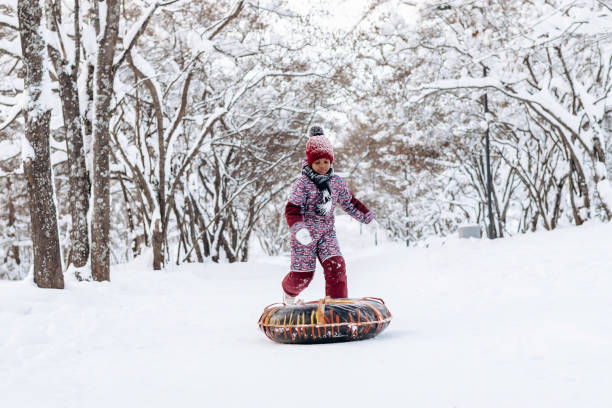 Happy little African-American girl in a red hat and jumpsuit rides on tubing in the winter park.Beautiful trees are covered with white snow.Winter fun,active lifestyle concept.