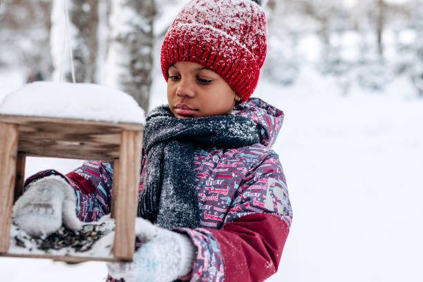 Little African-American girl in a red hat and jumpsuit pours food into a bird feeder in a winter forest.Taking care of animals.Eco-friendly concept