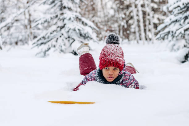Funny little African-American girl in a red hat and jumpsuit is lying in the snow.Winter fun,active lifestyle concept