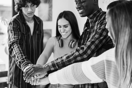 Multiracial young group of friends celebrating together stacking hands inside library room - Focus on hands - Black and white editing