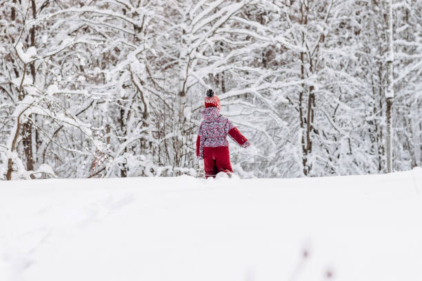 A little girl in a red hat and jumpsuit walks in the winter forest.Beautiful trees are covered with white snow.Winter fun,active lifestyle concept.