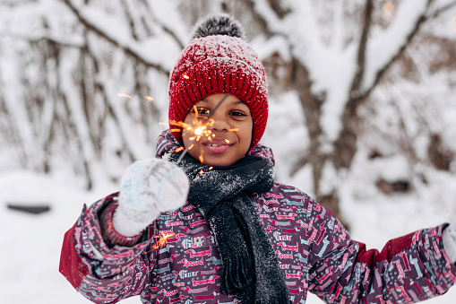 Happy little African-American girl in a red hat and jumpsuit holds a burning sparkler in her hands in a winter forest.Winter fun,merry Christmas and Happy New Year concept.