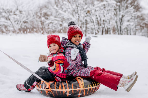 Happy Caucasian and African-American girls ride on tubing in the winter park.Beautiful trees are covered with white snow.Winter fun,active lifestyle concept