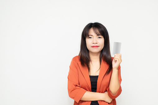 attractive asian business woman holding blank white mock up card in hand against white concrete wall background in office.