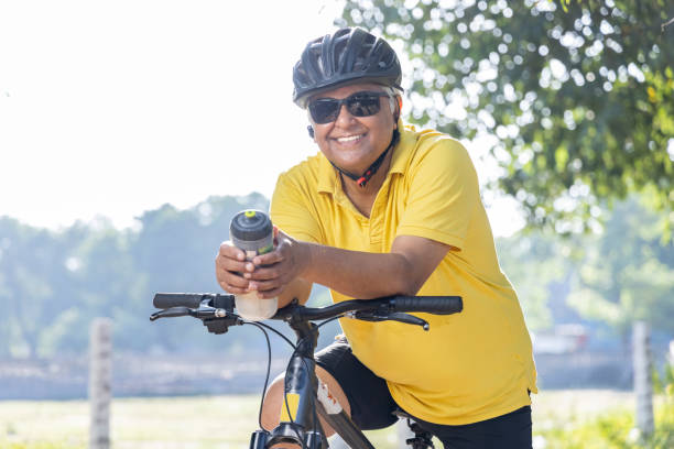 Happy senior man leaning on bicycle with water bottle Happy senior man in protective sportswear leaning on bicycle handlebar outdoors man 60s bicycle stock pictures, royalty-free photos & images