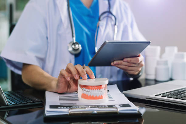 dentista concentrado sentado à mesa com amostras de mandíbula modelo de dente e trabalhando com tablet e laptop em consultório odontológico profissional clínica odontológica. - dental hygiene prosthetic equipment dentist office dental equipment - fotografias e filmes do acervo