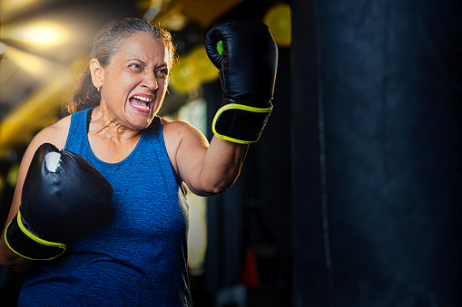 Aggressive old woman in sportswear training with boxing bag in gym