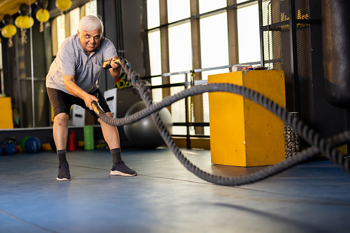 Active senior men in sportswear training with battle rope in gym gym