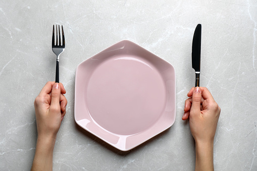 Woman with fork, knife and empty plate at light grey table, top view