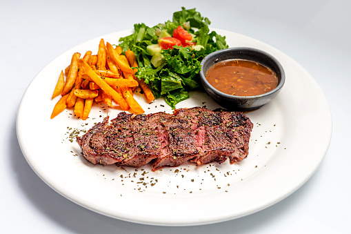 Gilled beef steak and potatoes served with pepper sauce and lettuce on black plate, top view