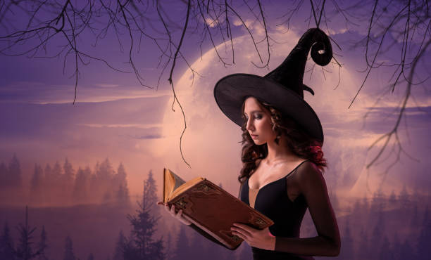 istock Halloween witch. A beautiful young woman in a witch's hat conjures, practices witchcraft with an old book in her hands against the backdrop of a huge lilac moon. Wide Halloween art design. 1427251856