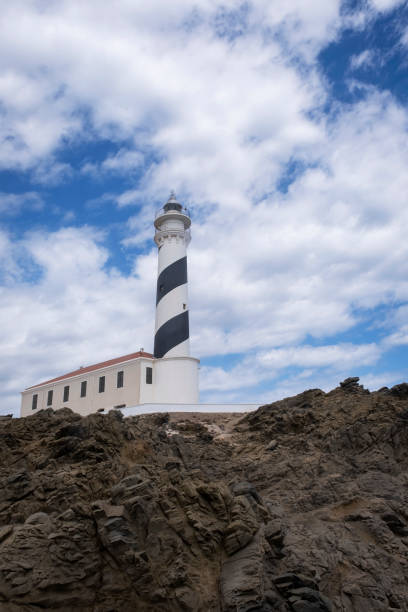 istock Favaritx lighthouse with its tower painted with a blue and white spiral on a rocky cliff, with the sky of cotton clouds, low point of view, copy space, horizontal 1427254056