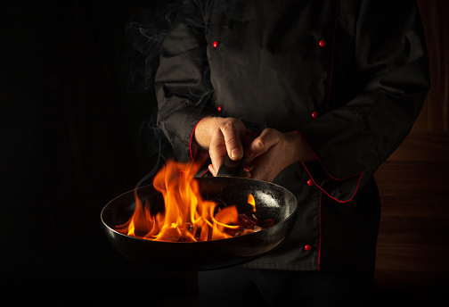 Professional chef cooking food in pan with fire flame on black background. Restaurant and hotel service concept. European cuisine.