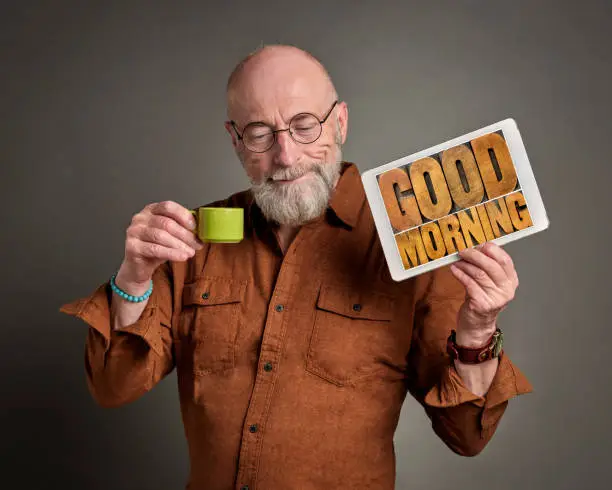 Good Morning - smiling senior man is drinking coffee and showing a digital tablet with text in wood type, positive start of  a new day concept