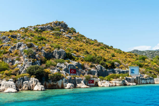 Hamidiye Bay in Kekova region of Antalya province of Turkey Hamidiye, Antalya, Turkey - August 14, 2021. Hamidiye Bay in Kekova region of Antalya province of Turkey, with Turkish flags and texts Hamidiye and How happy is the one who says I am a Turk! in Turkish written on the rocks. The bay has a historical feature as Hamidiye Battle Cruiser, which set sail from Canakkale on January 13, 1913 during the Balkan War and earned its captain-commander Huseyin Rauf Orbay the titles of The first captain-commander who executed a raid campaign in the history and Hero of Hamidiye by sailing 11.500 miles for 7 months and 24 days without any repair; anchored here  on the way to Aegean Sea from Beirut on the dates of January 25-26, 1913 for two days in order to rest, clean its boilers and trick the enemy. The height of the island created a natural and hidden shelter by preventing the Hamidiye Cruisers masts from being seen. kekova stock pictures, royalty-free photos & images