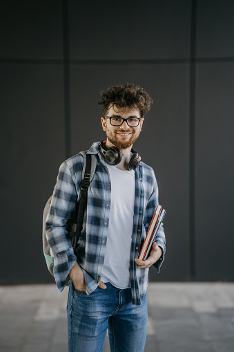 Portrait of male university student with eyeglasses, backpack and notebooks against gray wall of college building