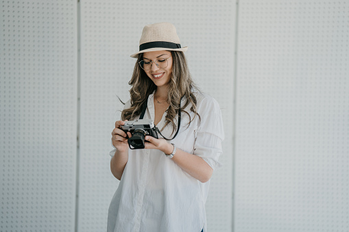 Beautiful young woman with retro camera standing against white wall on sunny day