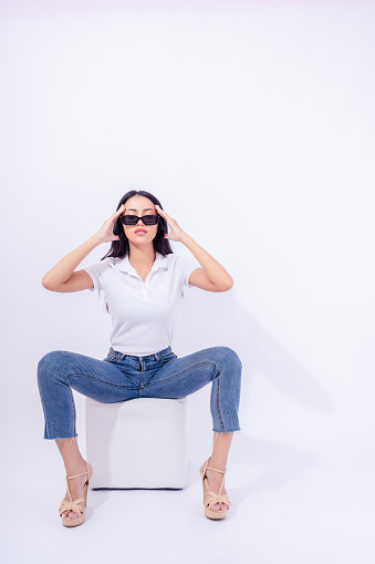 Beautiful young asian woman in Jeans High heel Collar shirt sitting on sofa cube isolated on white background in Studio Attractive girl wear sunglasses Full length of Asia female posing confident